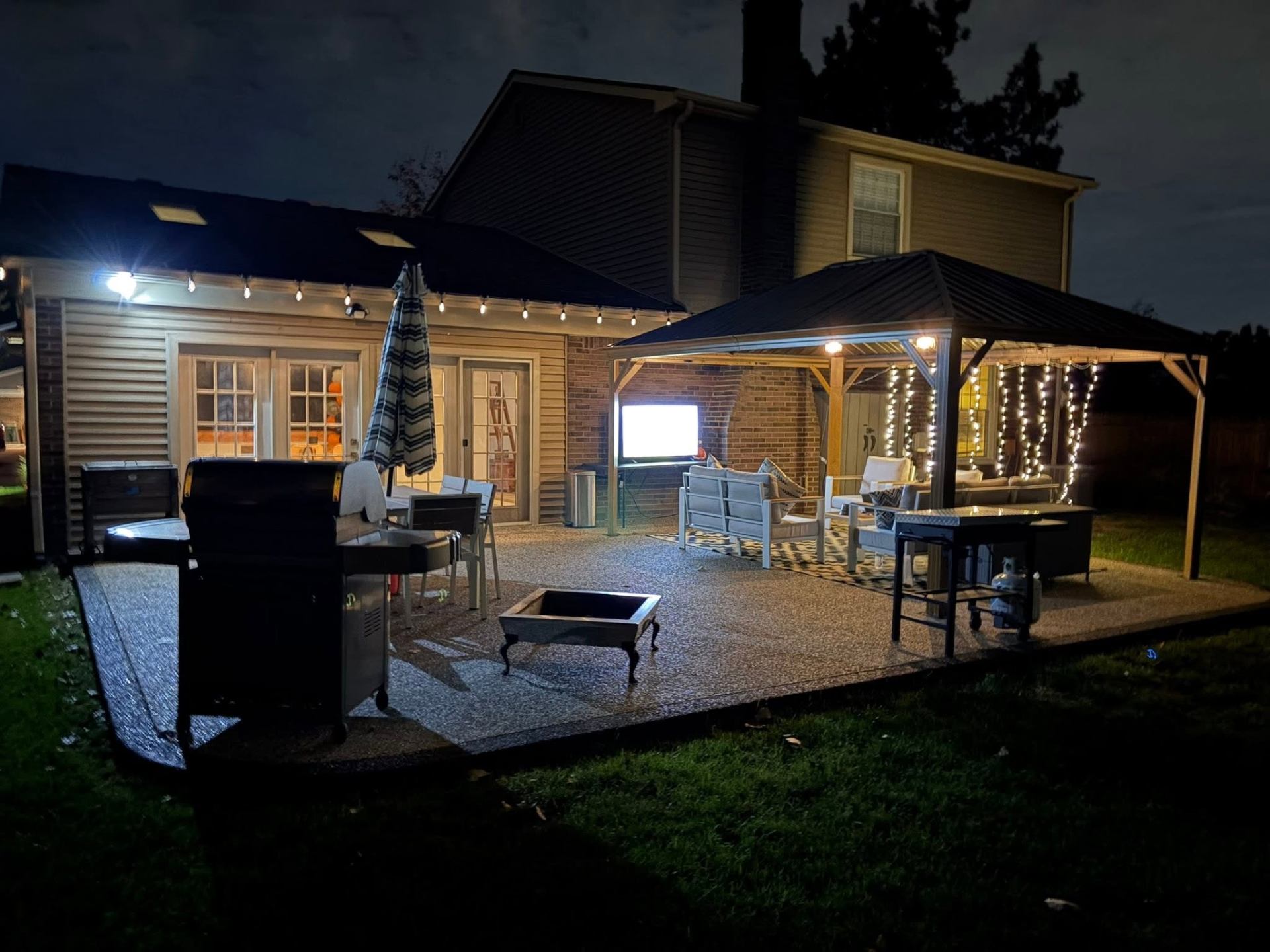Exposed aggregate patio with covered gazebo, grill, and dining area lit up at night