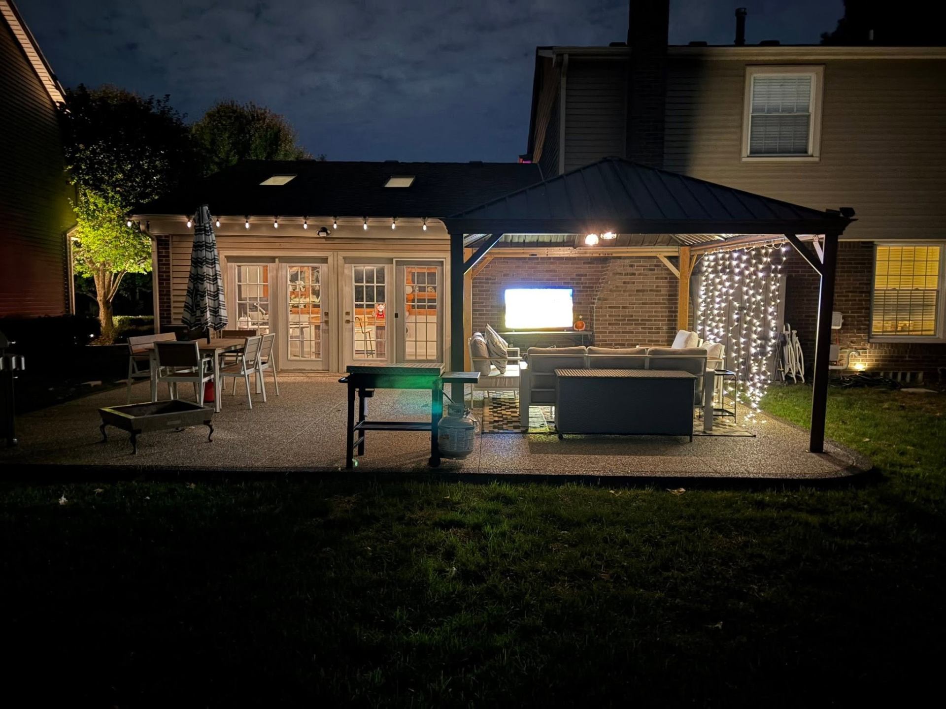 Backyard exposed aggregate patio at night with pergola, lounge seating, and string lights